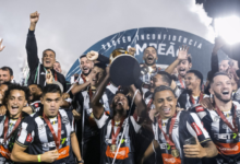 A imagem conta com diversos jogadores de futebol sorridentes. Eles vestem uma camisa preta e branca listrada, com uma medalha por cima do peito. No centro, um homem negro com uma faixa vermelha no braço (a faixa de capitão), levanta uma taça dourada, com sua base na cor preta virada para a câmera.