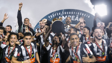 A imagem conta com diversos jogadores de futebol sorridentes. Eles vestem uma camisa preta e branca listrada, com uma medalha por cima do peito. No centro, um homem negro com uma faixa vermelha no braço (a faixa de capitão), levanta uma taça dourada, com sua base na cor preta virada para a câmera.