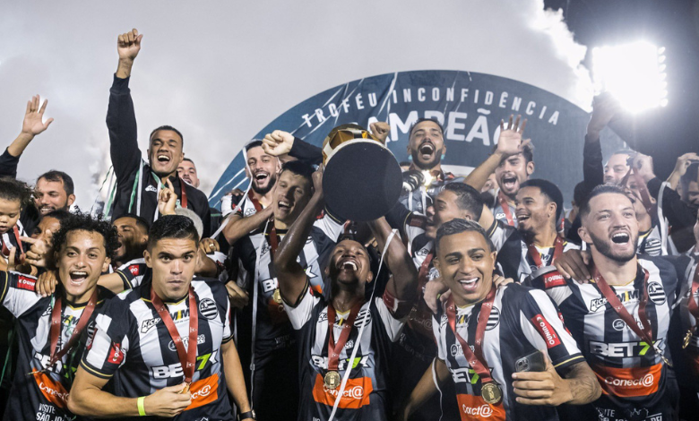 A imagem conta com diversos jogadores de futebol sorridentes. Eles vestem uma camisa preta e branca listrada, com uma medalha por cima do peito. No centro, um homem negro com uma faixa vermelha no braço (a faixa de capitão), levanta uma taça dourada, com sua base na cor preta virada para a câmera.