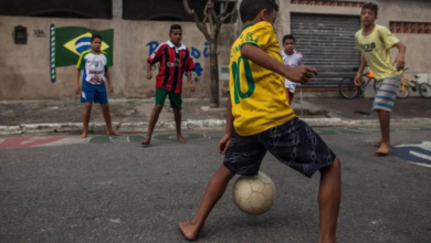 A foto mostra alguns meninos em uma rua. O garoto mais próximo a câmera está com a camisa amarela da seleção brasileira, com o número 10 em suas costas. Já em sua frente, está um menino com uma blusa com listras vermelhas e pretas, o uniforme do Milan da Itália. Também tem outros 2 garotos na cena. O primeiro menino está com uma bola, eles estão jogando o futebol de rua.