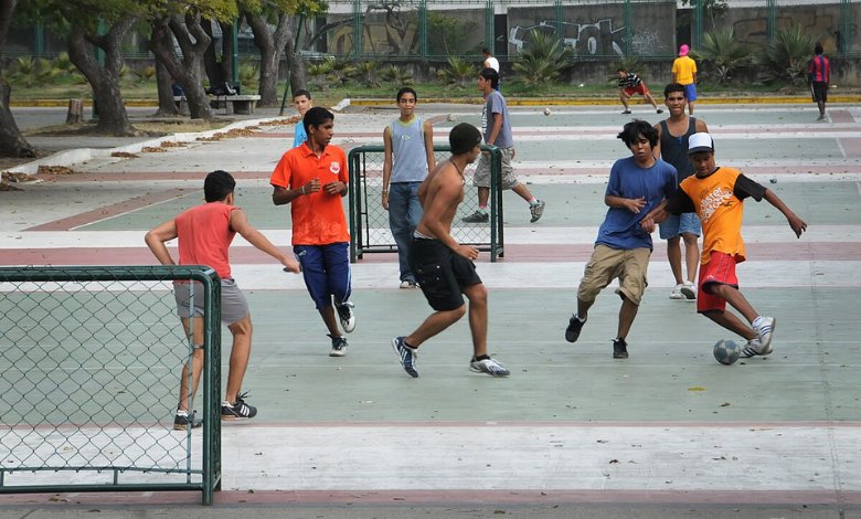 A foto mostra diversos adolescentes. Um deles, posicionado ao centro da imagem, está sem camisa. Os demais estão com suas camisas e bermudas longas. Todos eles estão em uma quadra de cimento,   se divertindo com uma pequena bola azul e branca. 