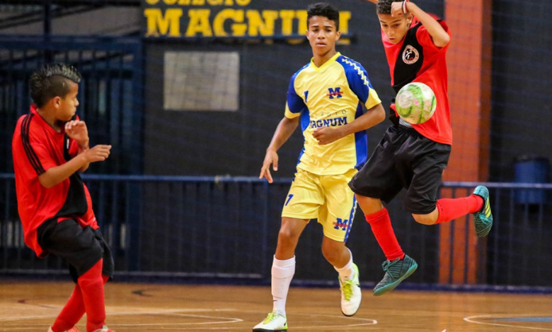 A foto mostra 3 garotos. 2 deles estão com uma blusa vermelha, com uma listra horizontal preta, shorts pretos e meiões vermelhos. O outro menino está com um uniforme amarelo, com detalhes azuis, shorts também amarelos com listras azuis e meiões brancos. Eles disputam os Jogos Escolares de Belo Horizonte, na modalidade Futsal.