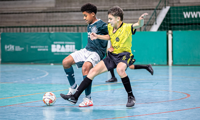 A imagem mostra um menino de camisa verde, shorts brancos e meiões verdes, disputando uma bola com outro garoto, esse de blusa amarela, shorts e meiões pretos. Os jovens estão em um campeonato de futsal sub-12.
