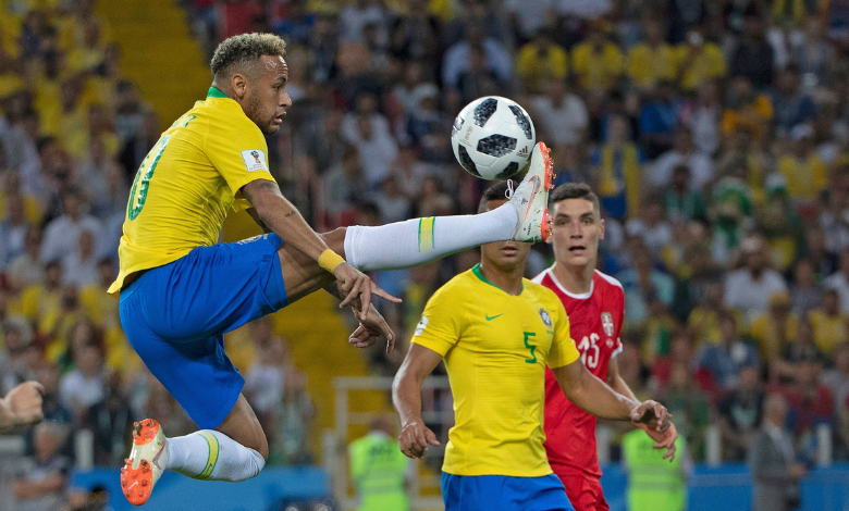 A foto mostra Neymar, na copa de 2018, dominando uma bola no ar, com a perna esticada na horizontal. Ao fundo, observando o lance, está o volante camisa 5, Casemiro. Ambos estão com o uniforme da seleção brasileira, uma camisa amarela, shorts azuis e meiões brancos. Eles se encontram em um estádio de futebol, que tem em sua torcida uma maioria de uniformes amarelos. 