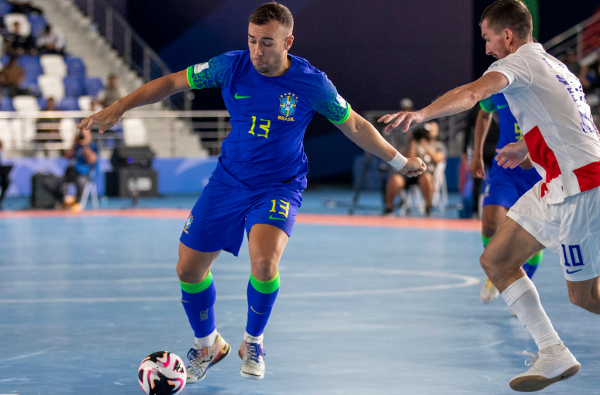 Na imagem vemos um homem branco, de blusas, shorts e meiões azuis com detalhes verdes. Ele está correndo com uma bola branca de detalhes pretos. Indo em sua direção está um homem branco, de blusas, shorts e meiões brancos de detalhes vermelhos e azuis. Esta é uma partida de futsal entre Brasil e Croácia.