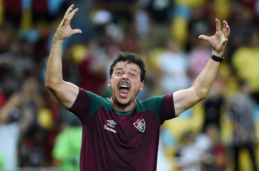 Na foto vemos Fernando Diniz, um homem branco, de barba preta e cabelo preto, vestindo uma blusa na cor vinho, com detalhes verdes em seus ombros. Este é o uniforme da comissão técnica do Fluminense. 